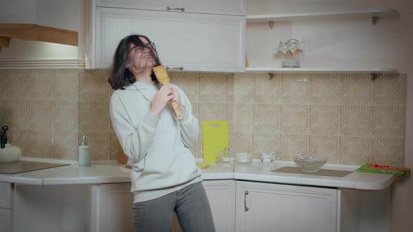 Girl in Glasses is Having Fun Dancing and Singing on the Background of the Kitchen Holding a Spoon alt