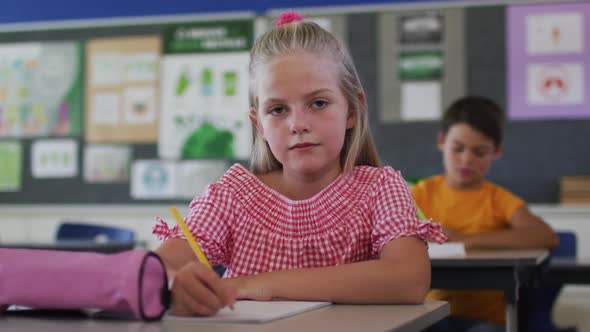 Portrait of happy caucasian schoolgirl sitting at classroom, making notes, looking at camera alt