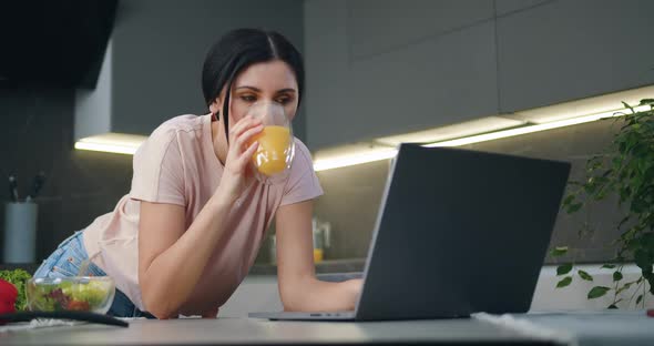 Brunette Working on Laptop which Standing on Kitchen Table and Drinking Orange Juice alt