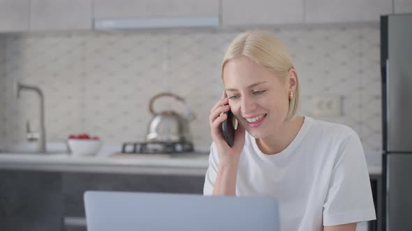 Young Blonde Woman Starts Phone Talk When Sitting at Laptop in Kitchen