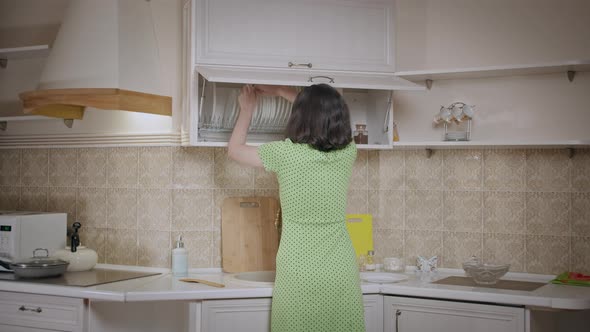 Girl in a Dress Puts Things in Order in the Kitchen Cabinet Arranges the Dishes Rear View alt