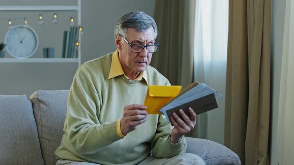 60s Elderly Man Recipient with Glasses Sorting Letters in Living Room Concentrated Mature alt