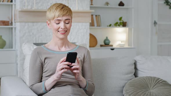 Relaxed Beautiful Woman Sitting on Couch Holding Smartphone Looking at Phone Screen Play Games Using alt