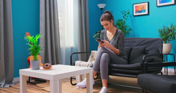 A Young Woman is Relaxing on the Living Room Couch Texting on Her Smartphone alt
