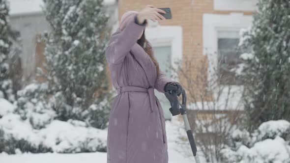 Happy Young Caucasian Woman Taking Selfie Outdoors Standing with Shovel on Snowy Backyard alt