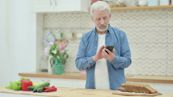 Old Man Using Smartphone While Cutting Vegetables in Kitchen alt