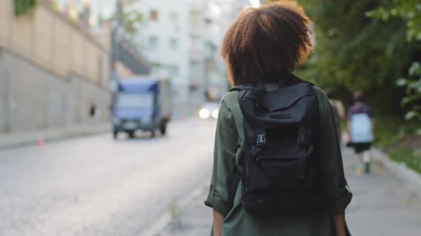 Back View Young African American Tourist with Afro Hair and Backpack Walking Alone Along Road alt