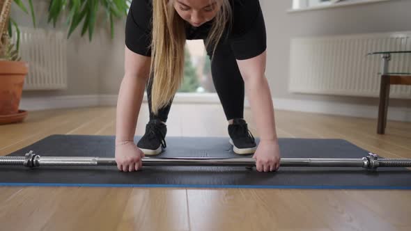 Chubby Caucasian Young Woman Raising Barbell Standing on Exercise Mat at Home alt