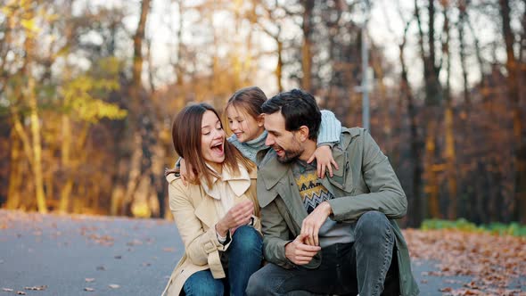 Young Smiling Parents Sitting Squatting in Autumn Park Little Daughter Running Up and Hugging Them alt