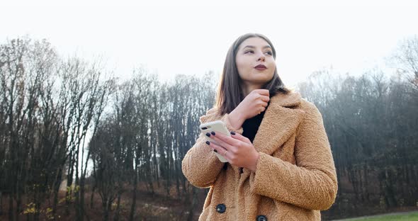 Portrait of Young Attractive Woman Smiling and Looking at Smartphone on the Green Lawn at Autumn alt