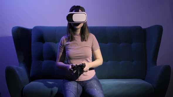 Woman Sits on Sofa in Virtual Reality Glasses Plays Video Game Using Gamepad