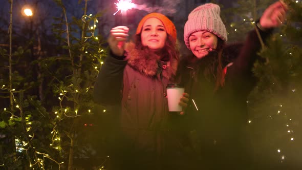 Two Young Smiling Women Holding Burning Sparkler and Coffee in Cold Winter Evening on Street