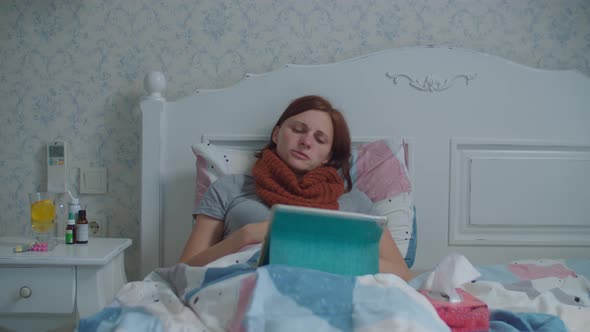 Sick Young Woman Using Tablet Computer Laying in Bed. Woman with Warm Scarf on Neck Coughs and Blows alt