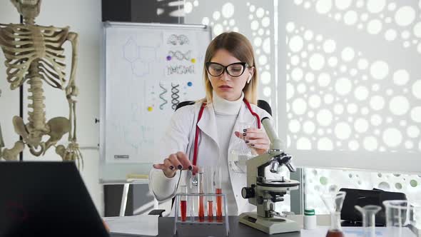 Female Doctor Sitting at the Table and Working with Test Tube and Flask with Chemical Solutions alt