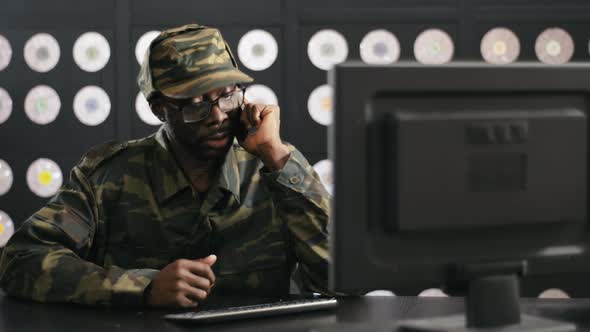 Bearded Black Man in Military Clothes Speaks on Phone and Looks at PC Display