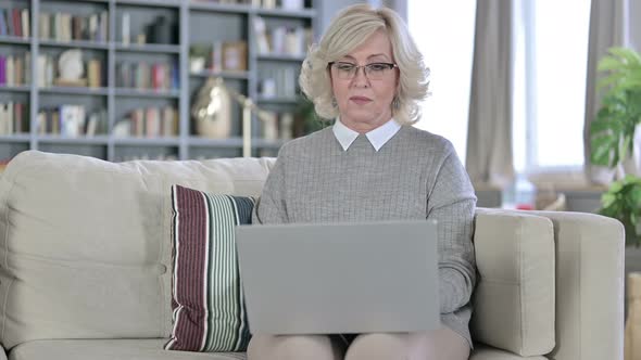 Serious Old Woman Sitting on Sofa and Using Laptop alt