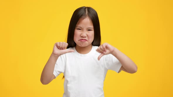 Naughty Little Asian Girl Gesturing Thumbs Down On Yellow Background alt