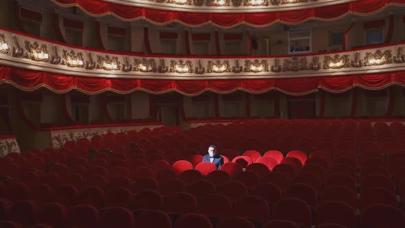 Rich man in mask in empty theater hall. Man spectator is sitting alone in large auditorium alt