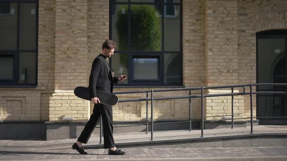 Side View Serious Successful Young Businessman Walking in Sunlight on City Street with Skateboard alt