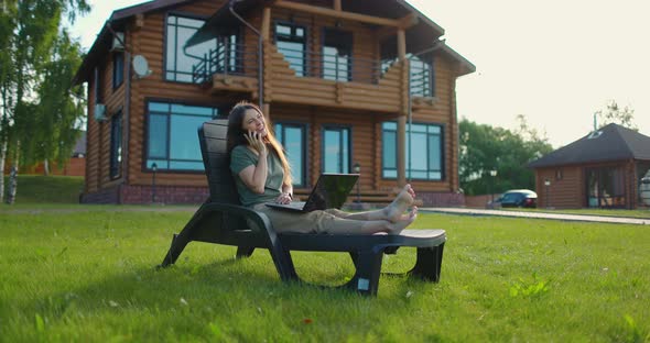 Young Happy Girl Sitting with Laptop in Lawn Chair at Backyard of Country House and Talking on alt