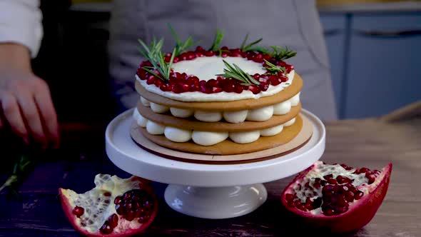 A cake consisting of three layers, cakes and white cream stands on a stand, decorated with pomegrana