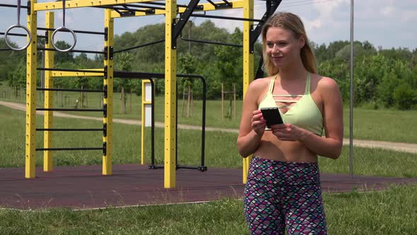 A Fit Beautiful Woman Works on a Smartphone at an Outdoor Gym alt