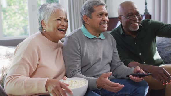 Happy biracial senior couple and african american friend watching tv with popcorn alt