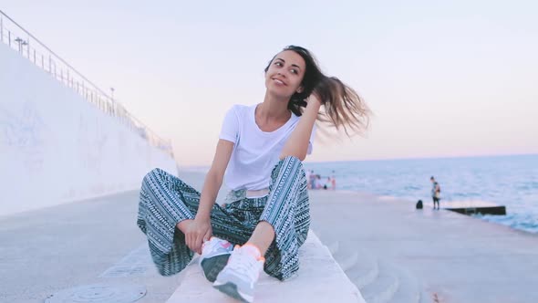 Beautiful Young Woman in Light Summer Clothes Sitting on Pier at Harbor at Dawn. Young Caucasian alt