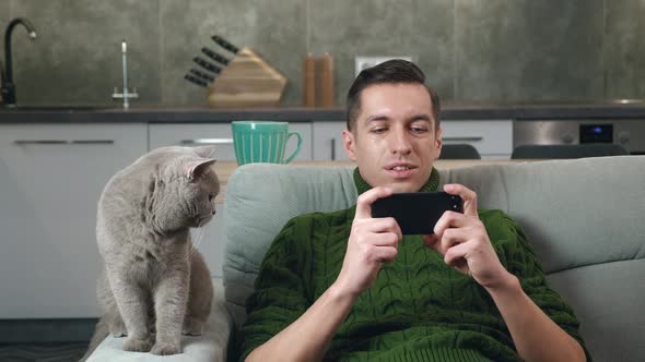 Young Man in Sweater Playing Application Games on His Smartphone While Resting on Sofa at Cosy Home alt