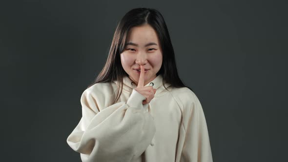 Smiling Asian Woman with Long Hair Holding Finger on Her Lips Over Grey alt