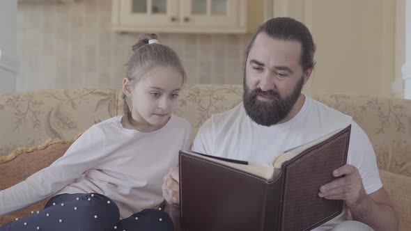 Portrait of Daughter and Her Father Sitting in Living Room and Looking Photo Album alt