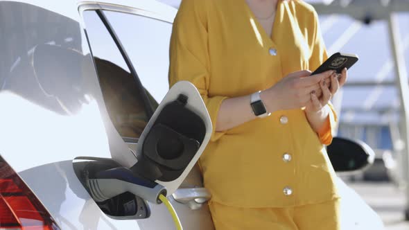 Woman Stands Near Recharging Electric Car With Mobile Phone