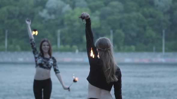Two Young Women Performing a Show with Flame Balls Standing on the Riverbank alt