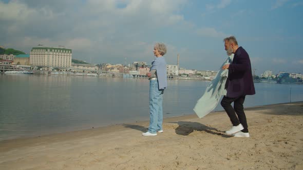 Senior Woman Standing Along River Bank and Man Covering Her with Plaid alt