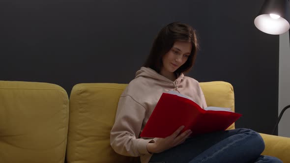 Attractive Longhaired Girl Sits at Home on Sofa and Smiling Reads Book