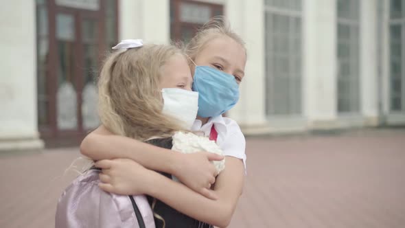 Close-up of Two Schoolgirls in Covid-19 Face Masks Hugging Outdoors. Portrait of Happy Caucasian alt