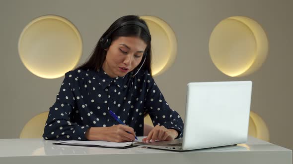 A Young Asian Woman Using a Headset Looks at a Laptop Screen Listens and Studies Online Courses alt