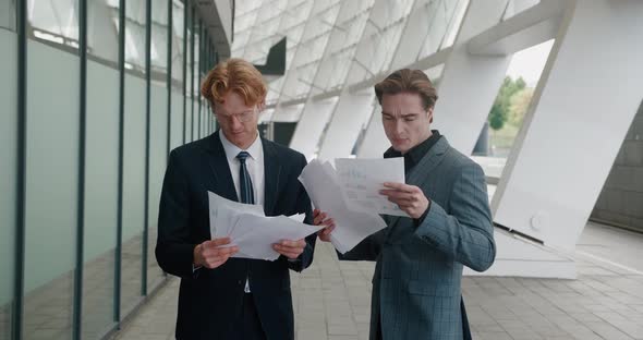 Two Young Businessmen in Formal Suits Holding Paper Douments in Hands Talking and Discussing alt
