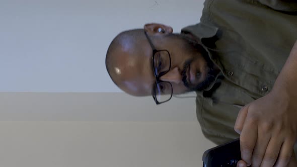 Bald Male Wearing Glasses Holding Smartphone Swivelling In Chair. Vertical Video, Static Shot alt