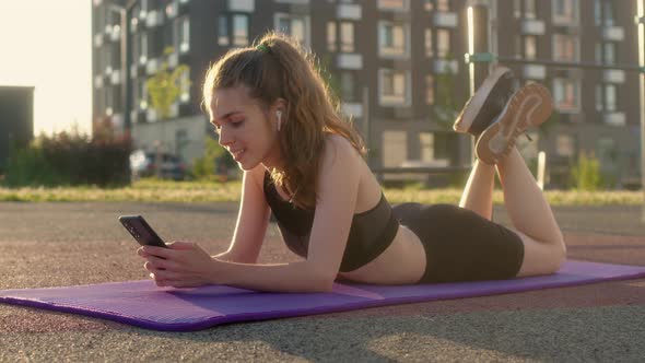 Cheerful Happy Young Woman Lies on Fitness Mat During Break Between Outdoor Sports and Communicates alt