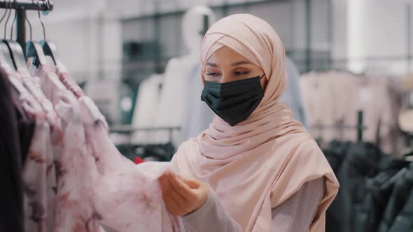 Young Arab Woman Customer Buyer in Hijab Wearing Protective Medical Mask in Clothing Store Carefully alt