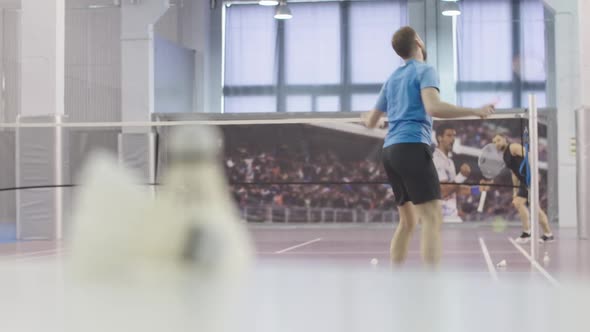 Wide Shot of Two Men Playing Badminton on Indoor Court. Rack Focus Changes To Shuttlecock Lying at alt