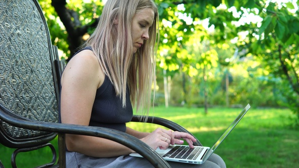 Beautiful blonde girl typing on a laptop while sitting in a rocking chair outdoors