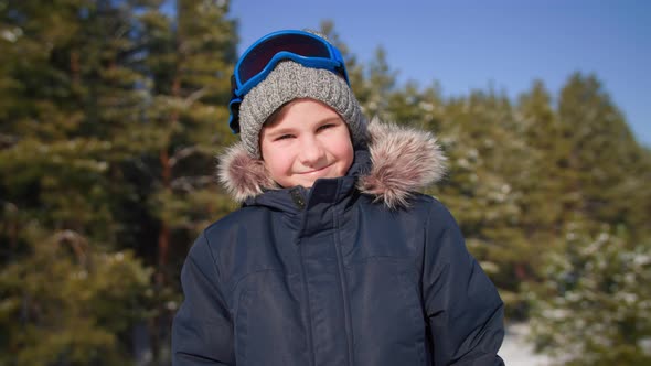 Portrait of Cute Male Child in Ski Goggles and a Hat Stands Background of Trees in a Winter Forest