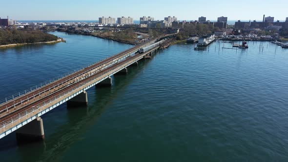 An aerial shot over Grassy Bay in Queens, NY. The camera dolly in behind a train on elevated tracks alt