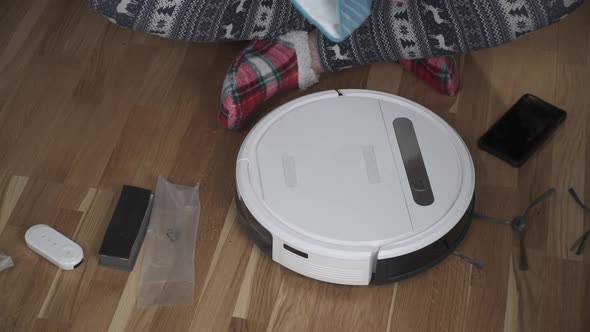 A Young Father in Sleepwear with a Winter Ornament and Christmas Funny Socks Serves a Roobot Vacuum alt