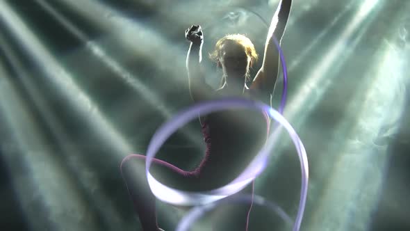 Against a Smoky Background, a Young Sportswoman Actively Trains with a Silk Ribbon in the Spotlight alt