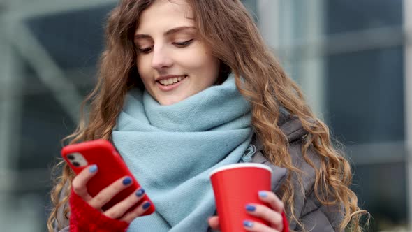 Young Girl is Laughing and Reading Something on Her Smartphone While Holding a Cup of Coffee