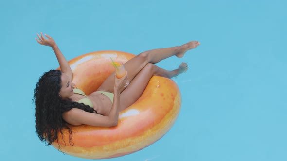 Attractive Slim Girl Model with Long Tanned Legs Resting Drinks Cocktail Lies in Orange Inflatable alt