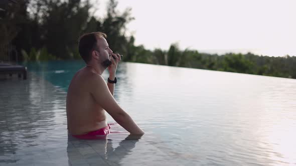Adult Man Talking on Phone Enjoying Sunset View From Infinity Pool in Rooftop Tropical Hotel alt